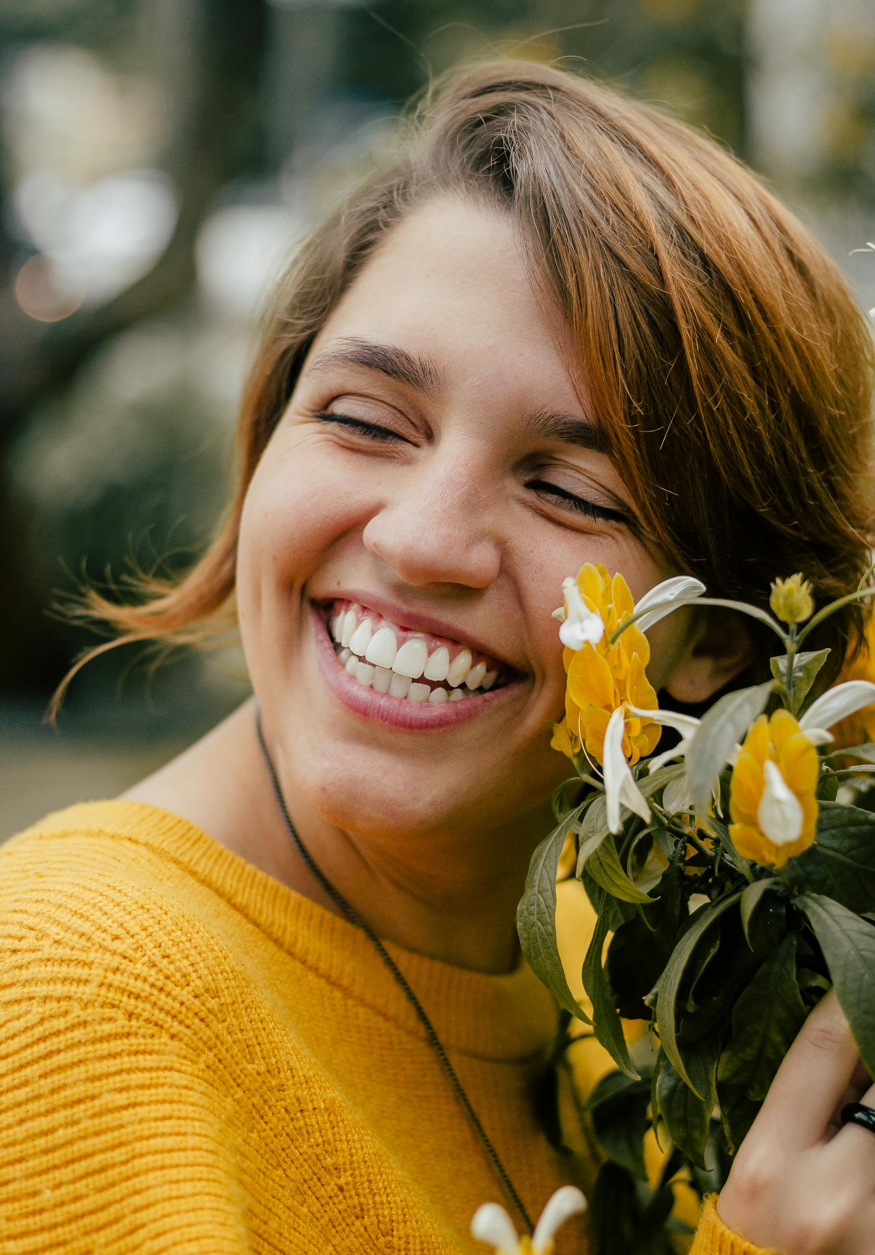 A smiling woman enjoys a sunny day outdoors holding bright yellow flowers.